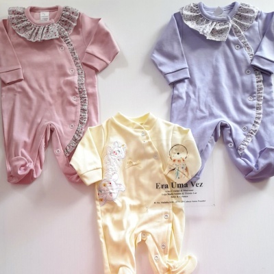 Três macacões de bebé rosa, amarelo e lilás com detalhes florais e bordados, e texto de marca no amarelo.