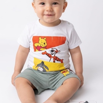 Bebé com t-shirt branca estampada Ursinho Pooh e calções verdes sentado no chão branco com cartão ao lado.