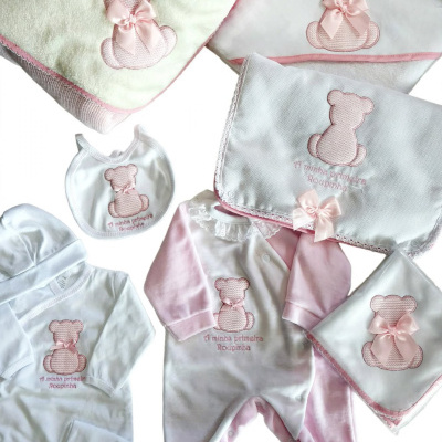 Roupas e acessórios de bebé em branco e rosa com ursinho bordado e laço.