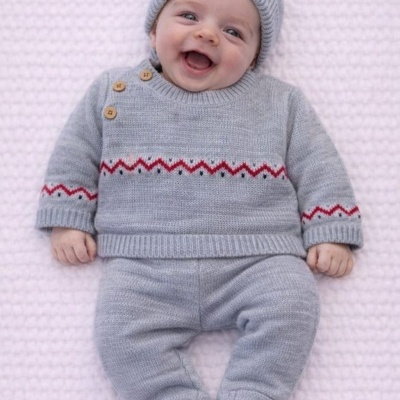 Bebé sorridente deitado com conjunto de malha cinza com padrão vermelho e preto e gorro, sobre manta rosa claro.