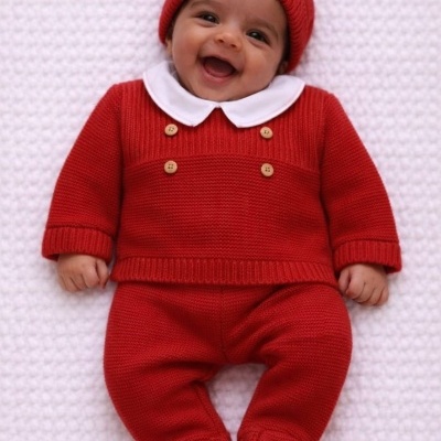 Bebé vestido com roupa vermelha de malha com gola branca e botões, deitado sobre superfície branca.