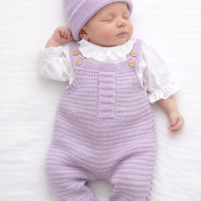 Bebé a dormir vestido com macacão e gorro lilás de lã e camisa branca numa superfície branca