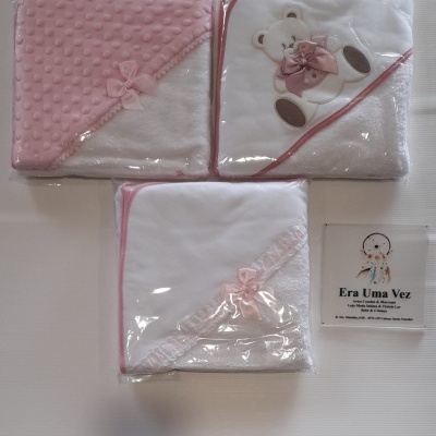 Três toalhas de bebé cor-de-rosa e branca embaladas em plástico, uma com ursinho bordado e laço, outras com detalhes em rosa e renda.