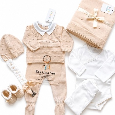 Conjunto de roupa de bebé bege e branco com malha fina, gorro, sapatos e manta enrolada.