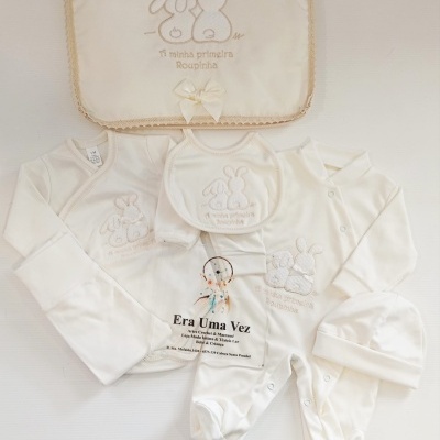 Conjunto de roupa de bebé branco com bordados de coelhos e texto, manta com renda e laço