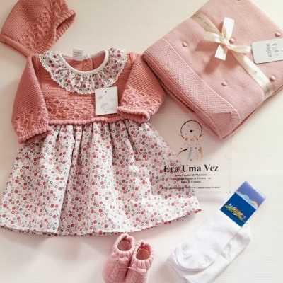Conjunto de roupa de bebé com vestido floral, touca, sapatinhos, meias brancas e manta rosa tudo disposto sobre fundo branco