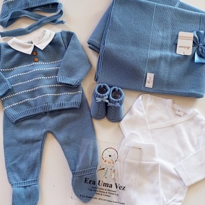 Conjunto de roupa de bebé azul e branco com detalhes tricotados e body branco sobre superfície clara