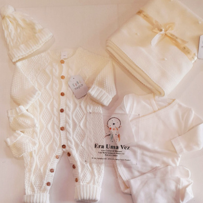 Conjunto de roupa de bebé branca em malha com gorro, macacão com botões e manta dobrada.