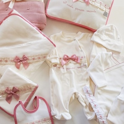 Conjunto de roupa de bebé branco com detalhes rosa, incluindo macacão, gorro, camisola, babete, manta, toalha e estojo, com laços rosa e renda.