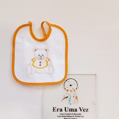Babete branco com bordas laranja e ursinho bordado junto de placa informativa da loja