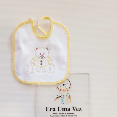 Babete branco com bordas amarelas e ursinho bordado colocado sobre uma placa com texto