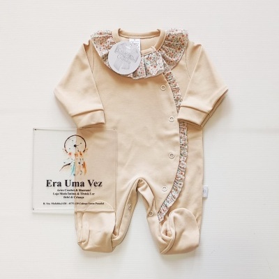 Macacão bege para bebé com folho floral e botões de pressão, sobre fundo branco texturado