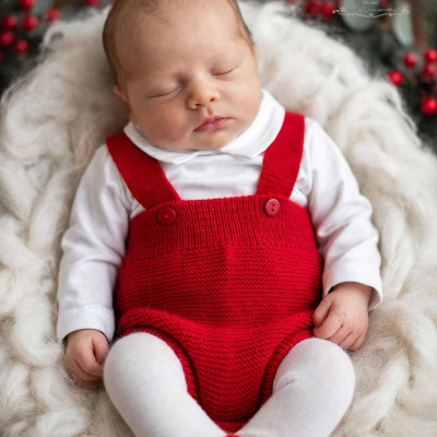 Bebé vestido com camisola branca, camisola vermelha de malha e botins vermelhos a dormir num fundo branco macio com decoração natalícia.