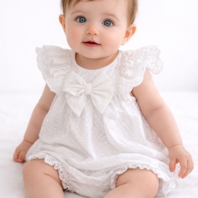 Bebé com macacão branco de bordados e laço sentado em fundo branco