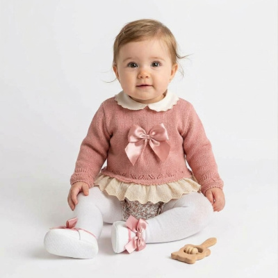 Bebé com camisola rosa, collants brancos e sapatinhos com laço rosa sentada ao lado de brinquedo de madeira.