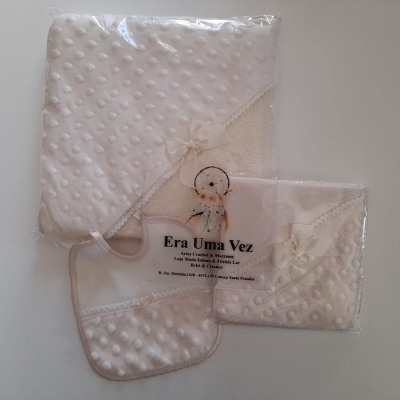 Conjunto branco para bebé com manta, babete e toalha com padrão de bolinhas em relevo e laço, embalados em plástico.