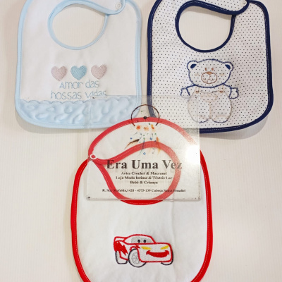 Três babetes de bebé com desenhos bordados e diferentes cores de borda sobre fundo branco.