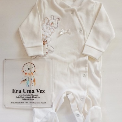 Pijama branco de bebé com bordado e laço e botões de pressão ao lado de placa com texto