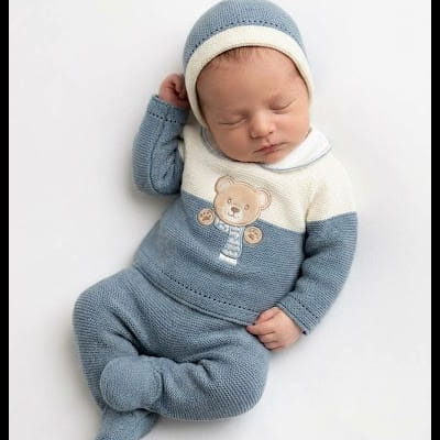 Bebé com macacão de malha azul e branca com urso bordado e gorro