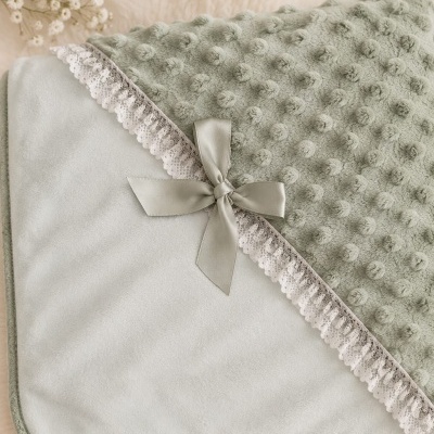 Manta de bebé verde com detalhes em renda branca e laço, sobre superfície creme com flores, coração e brinquedo de urso.