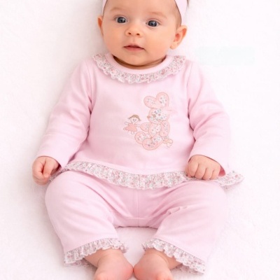 Bebé com conjunto rosa claro com bordados e renda florescida, e banda de cabelo com laço rosa.