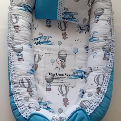 Cama de bebé com padrão de balões, aviões e animais, bordas almofadadas, laço azul, e almofada azul.