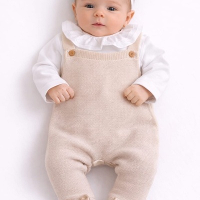 Bebé com macacão bege e fita branca com laço na cabeça deitada sobre fundo branco