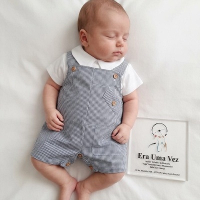 Bebé dormindo com jardineira quadrados azul e branco com botões, camisa branca, placa rústica ao lado