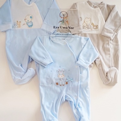 Três macacões de bebé azul claro e beige com bordados, em superfície clara, com placa de acrílico no meio.