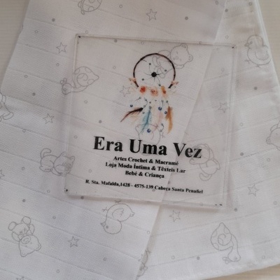 Fralda branca de algodão com padrão de animais e estrelas e etiqueta transparente com texto em português.