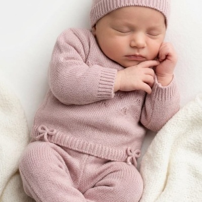 Bebé vestido com fato e gorro de malha rosa claro a dormir sobre manta branca.