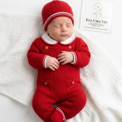 Bebé deitado com roupa vermelha de malha e gorro, manta branca e etiqueta à direita