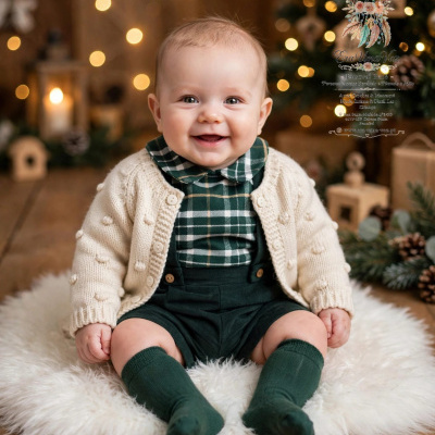 Bebé sorridente vestido com camisola creme e camisa xadrez sentado numa almofada branca com decoração natalícia ao fundo