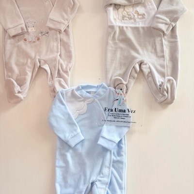 Três pijamas de bebé com manga comprida em tons neutros e azul claro, com bordados e botões frontais