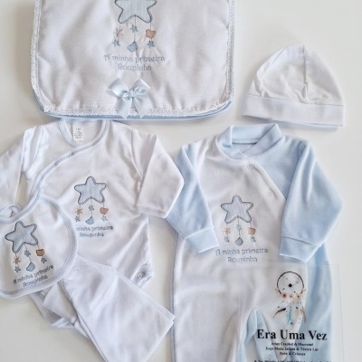 Conjunto de roupa de bebé branco e azul claro com decoração de estrela, texto e saco com laço azul.