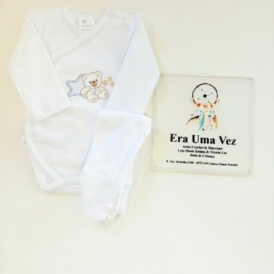 Body de bebé branco com bordado de ursinho e estrela azul, ao lado placa com texto informativo da loja.
