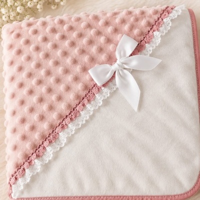 Manta para bebé cor de rosa e branca com fita e renda, ursinho de croché e flores brancas ao fundo.