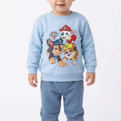 Camisola azul claro com personagens da Paw Patrol e calças azuis escuras em menino de pé sobre fundo branco