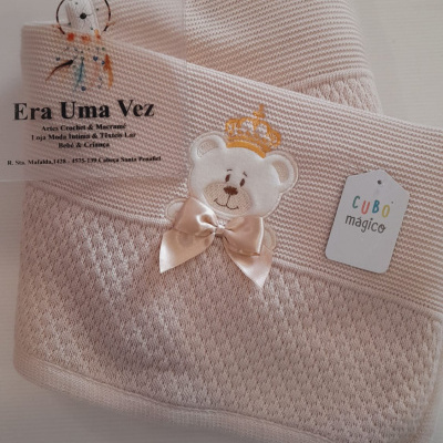 Manta infantil creme com urso bordado, laço cetim e etiquetas, sobre fundo branco.
