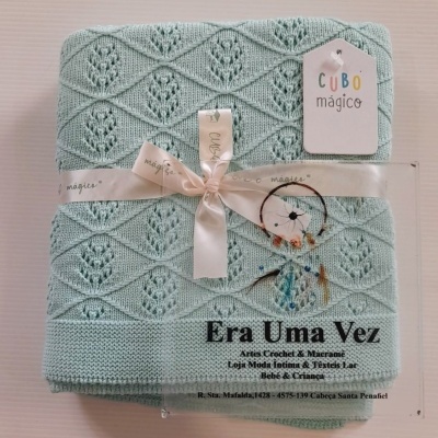 Manta azul-clara com padrão trançado, fita creme com texto e etiqueta pendente com texto