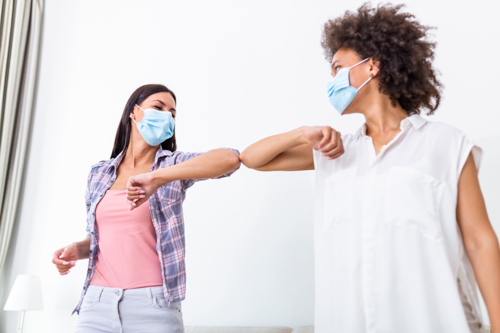 Duas mulheres com máscaras azuis cumprimentam-se com o cotovelo numa sala branca