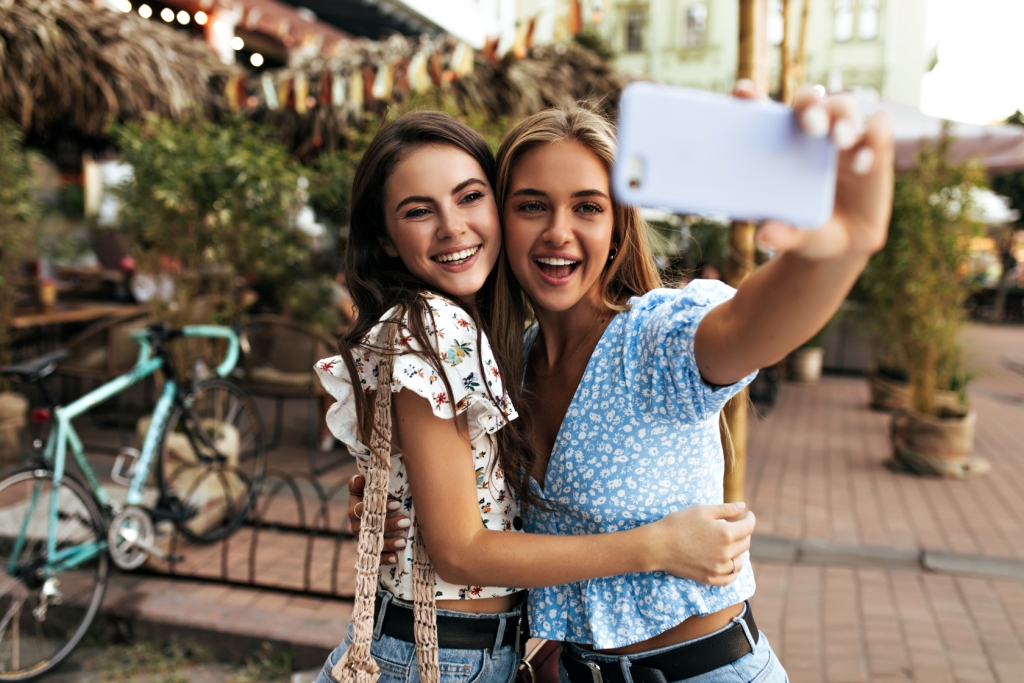 Duas jovens mulheres a tirar uma selfie na rua vestem blusas florais, com bicicletas e árvores ao fundo.