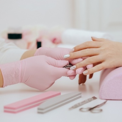 Manicure com luvas rosa a tratar unhas de cliente com apoio rosa e ferramentas visíveis na mesa.