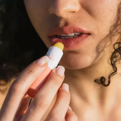 Baton labial com embalagem branca e cor de pêssego sendo aplicado nos lábios de uma pessoa.