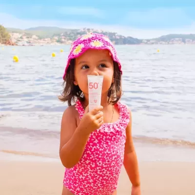 Criança com fato de banho e chapéu rosa segurando protetor solar 50+ na praia