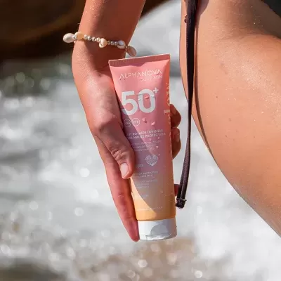 tubo de protetor solar SPF 50+ Alphanova Daily Sun na mão de mulher com pulseira, fundo de água