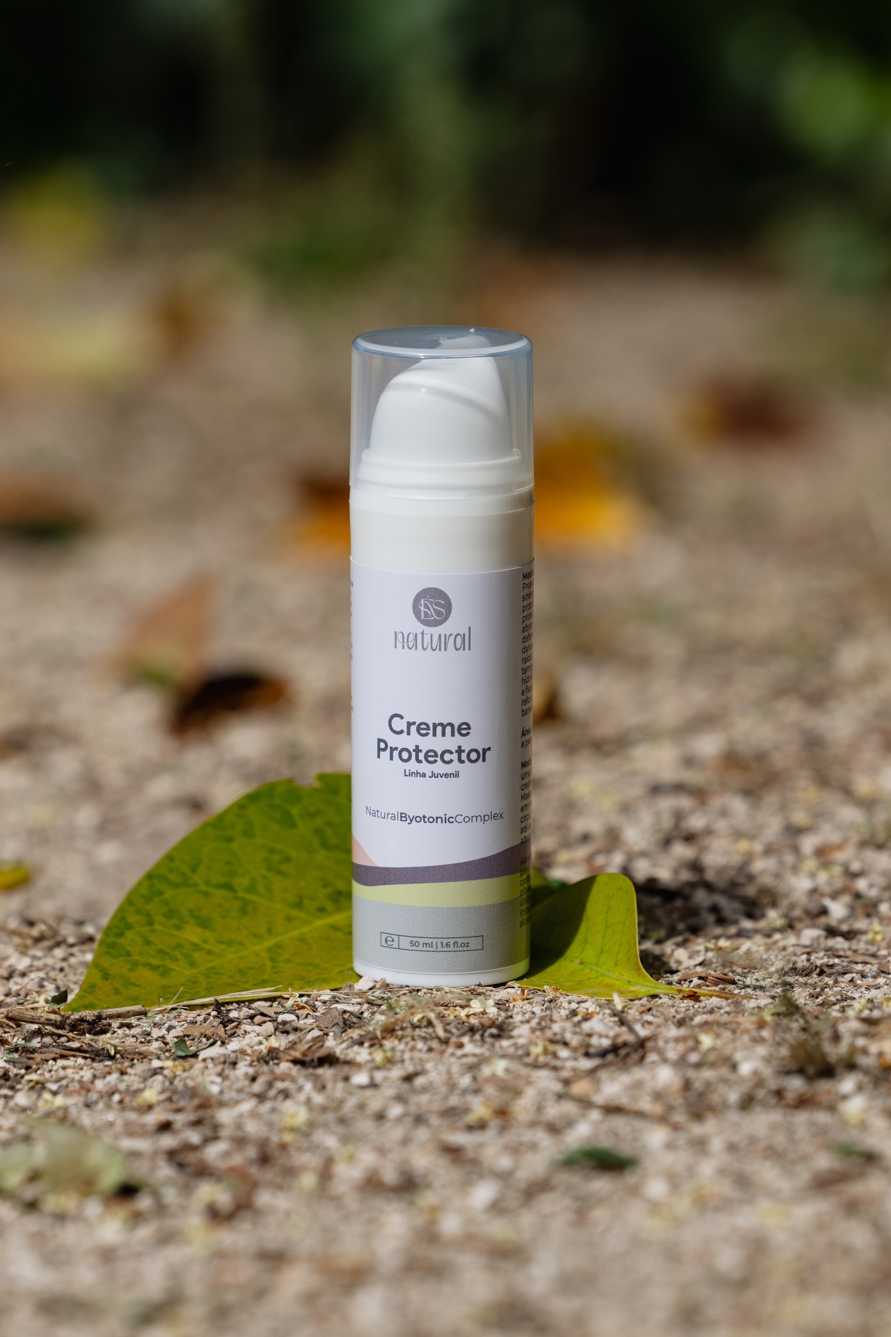 Creme Protector Solar FPS 50+ Frasco branco de creme protetor com tampa transparente sobre folha verde no chão de cascalho