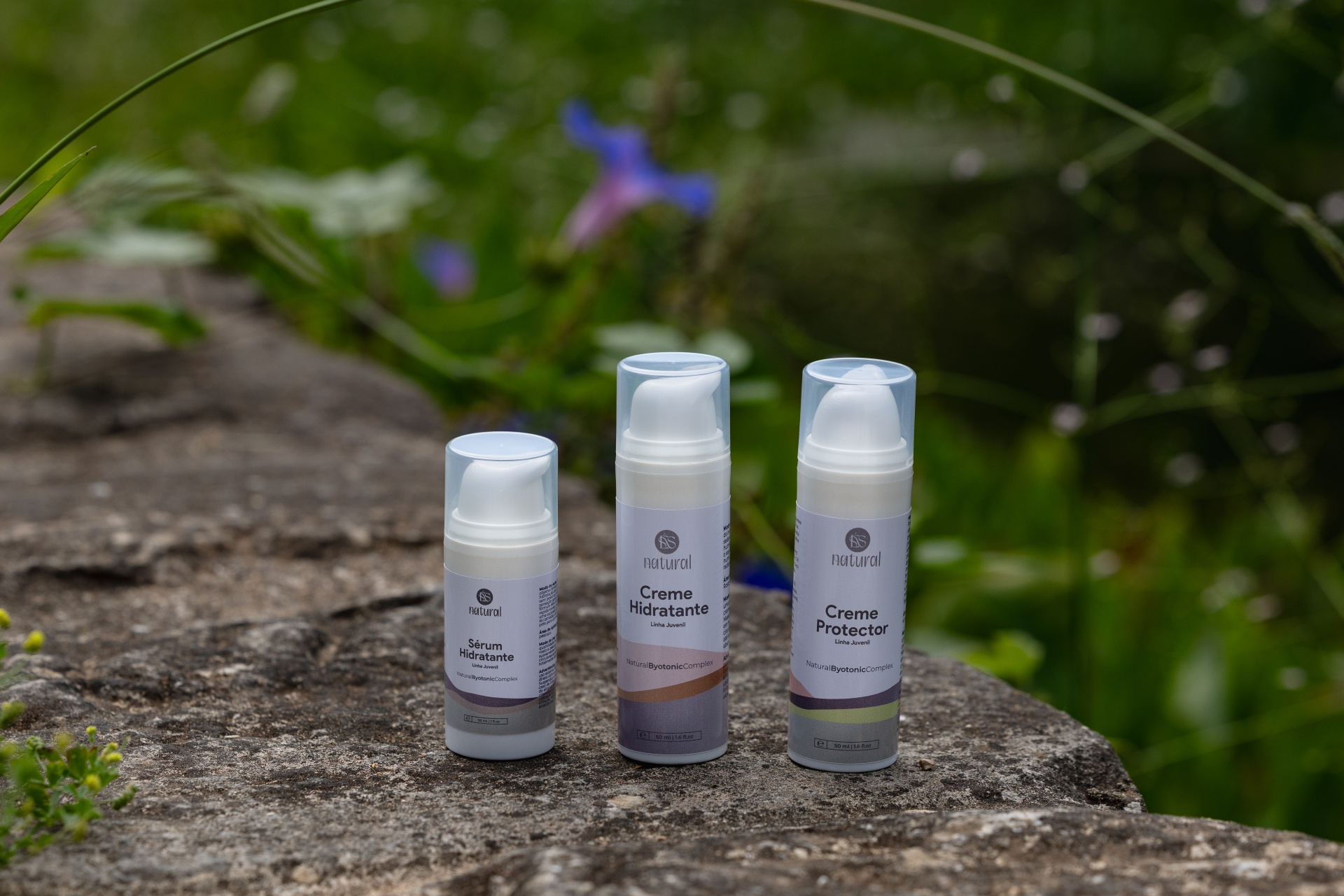 Linha Juvenil (inclui 3 produtos) Três frascos de creme cosmético sobre parede de pedra com plantas ao fundo