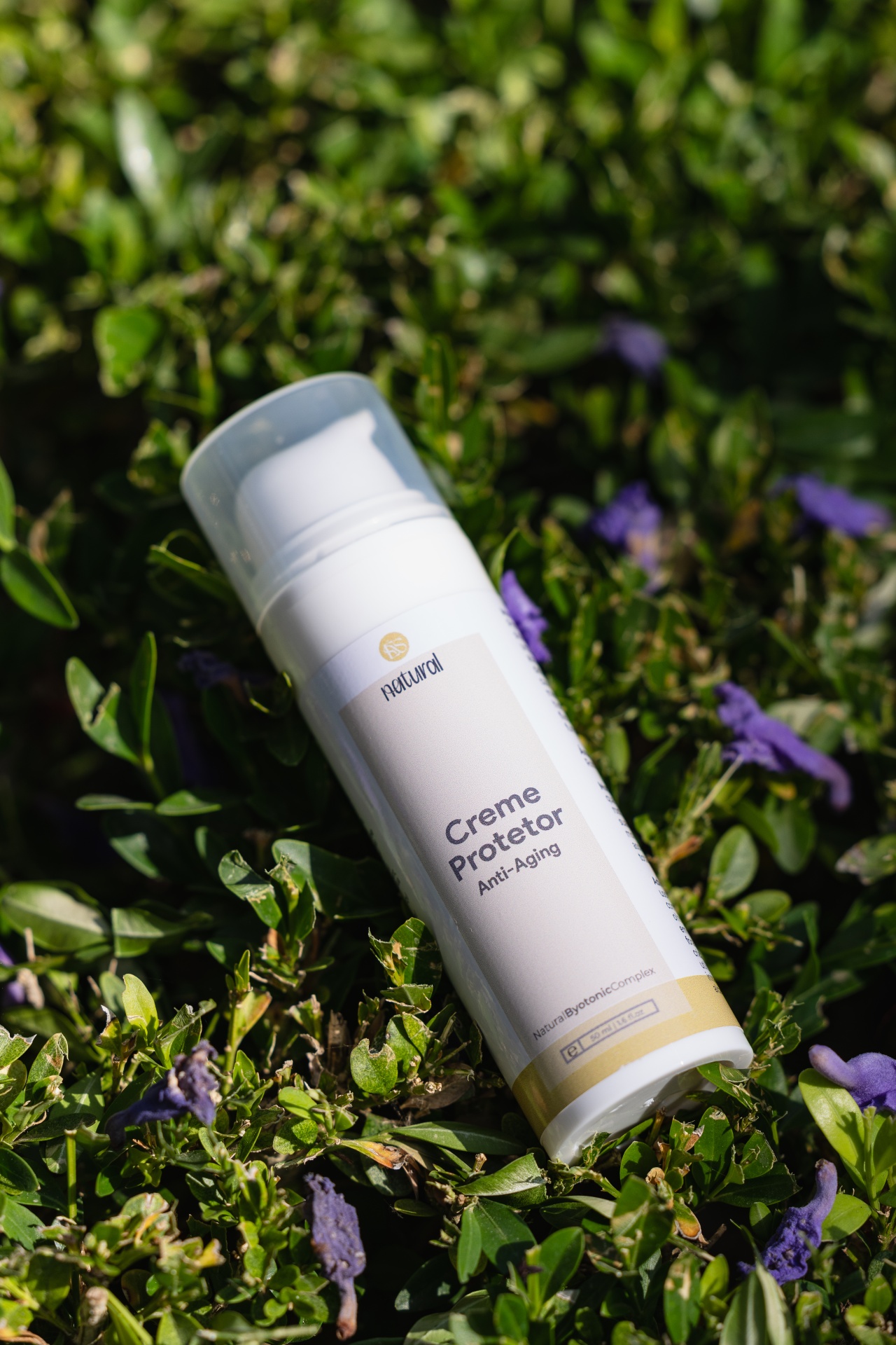 Creme Protetor Solar Anti-Aging FPS 50+ Frasco branco de creme protetor anti-envelhecimento em plantas verdes com flores roxas