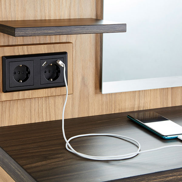 Tomada elétrica dupla com cabo branco ligado na parede de madeira clara junto a telemóvel sobre superfície escura